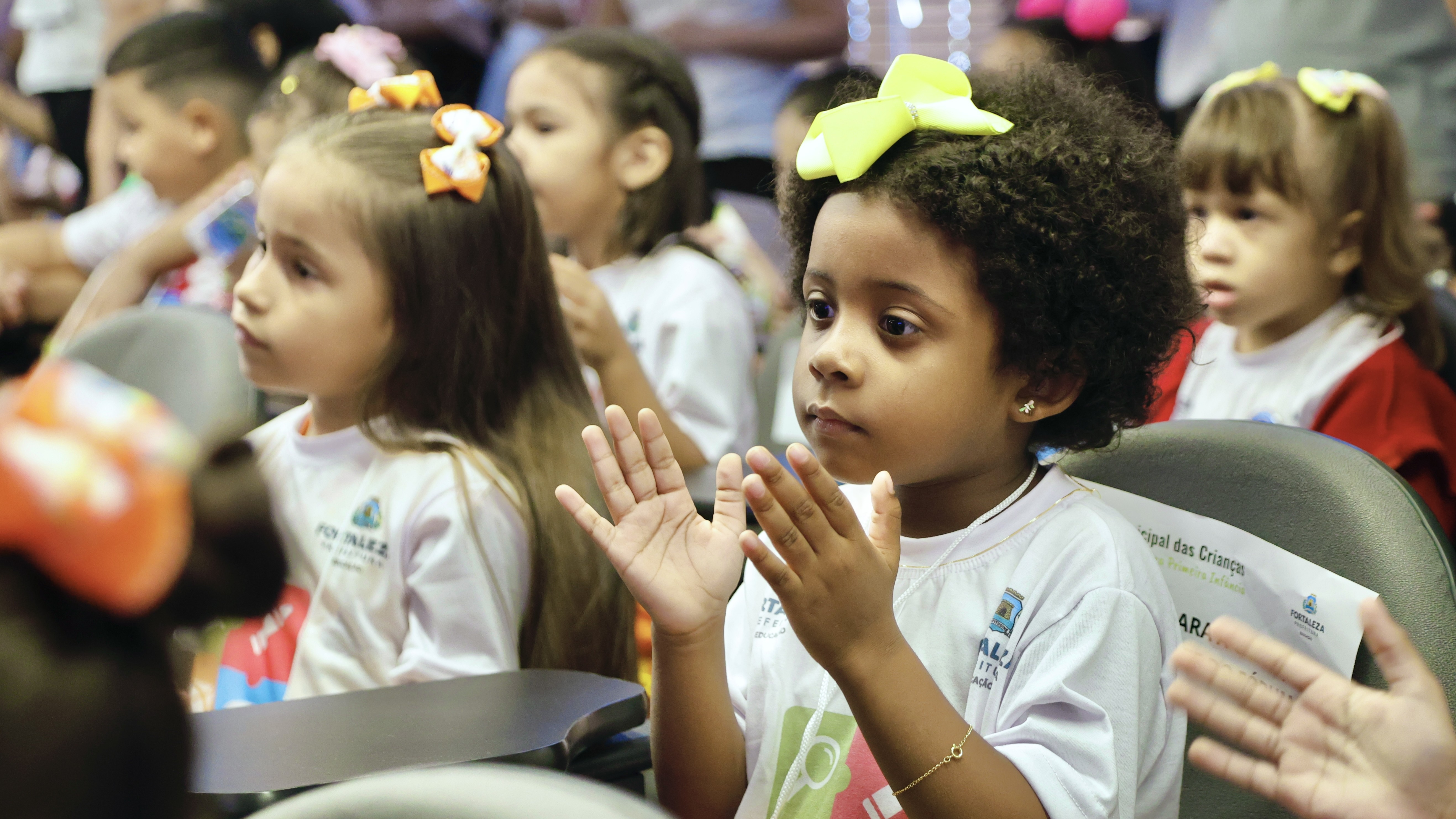 duas crianças, uma branca e uma negra, ambas de camiseta branca sentadas em cadeiras no auditório